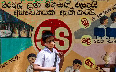 A schoolboy wearing a face mask and uniform walks past a colourful Sinhala-language mural featuring illustrated students.