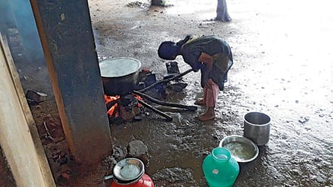 குடிநீர் தொட்டிக்கு கீழே விறகு அடுப்பில் சமைத்தவற்றை மழையில் நனைந்த வாறே எடுத்துச் செல்லும் பணியாளர்.