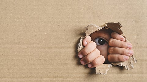 A child's eye and hands peer through a torn box.