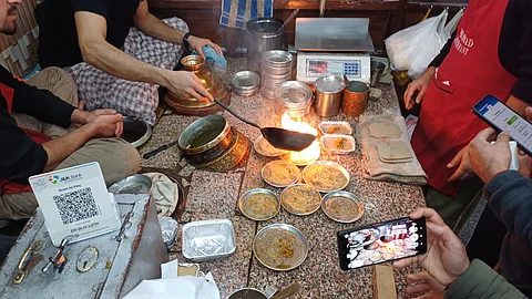 The shop owner pours hot oil over Harisa as curious customers wait, with one capturing the moment on their phone.