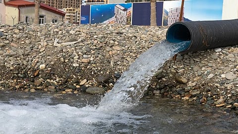 A large black industrial pipe discharging a steady stream of water onto a rocky bank.