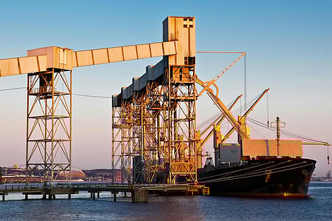 A large cargo ship docked at a marine industrial facility featuring tall metal loading structures and conveyor belts under a clear sky.