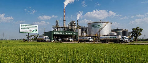 A wide shot of an "Ethanol Blending Plant" in India, featuring large storage tanks and industrial chimneys under a blue sky, directly adjacent to lush green agricultural fields.