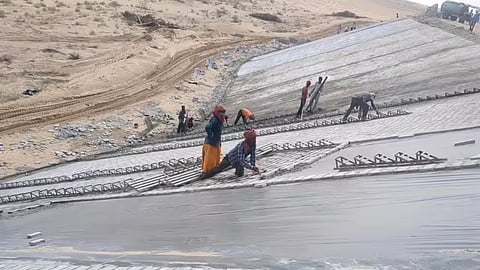 onstruction workers manually installing heavy-duty lining sheets and structural grids on a wide, slanted concrete embankment in a sandy desert region