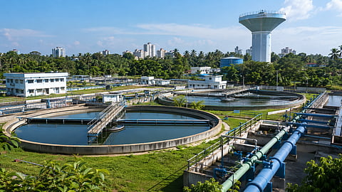 Modern water treatment plant with circular treatment tanks, large pipelines, utility buildings and an elevated water storage tower in a tropical urban setting.