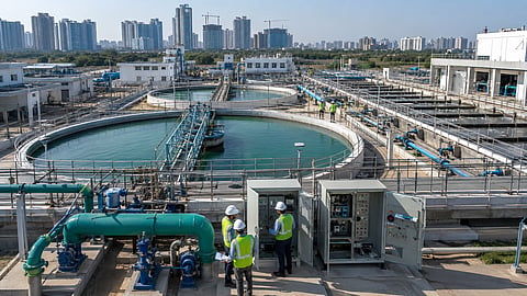 Engineers inspect electrical control panels and pumping equipment at a large water-treatment plant with circular clarifiers, filtration units, pipelines and a high-rise city skyline in the background.
