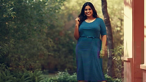 A confident plus-size Indian woman wearing a well-fitted midi dress featuring thin, evenly spaced horizontal stripes. She is posing outdoors in natural light, smiling, accessorized with simple jewelry and is comfortable.