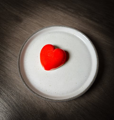 A heart-shaped red dessert, the Benami Kheer, presented on a minimalist white plate with a grey rim