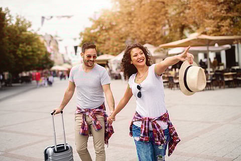Smiling cute mature couple holding hands and walking on the street on springtime. Woman pointing with finger while man carrying luggage.