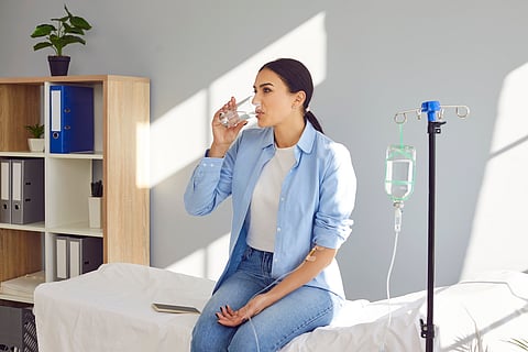 Young woman with sterile needle in arm sitting on medical bed by pole with IV drip bottle, drinking glass of water and receiving vitamin therapy infusion to boost immune system and suppress infection