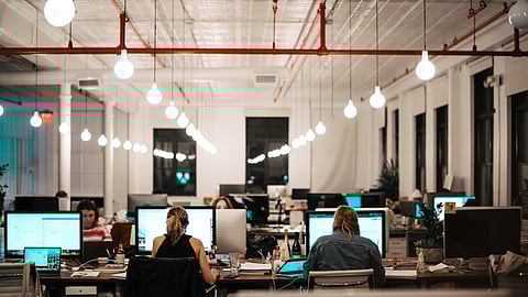 Group of individuals engaged in work at desks with computers, showcasing a busy office atmosphere.