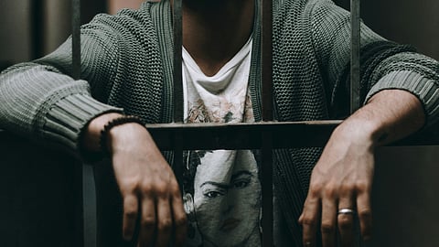 A man sits behind bars in a jail cell, looking contemplative and isolated in a dimly lit environment.