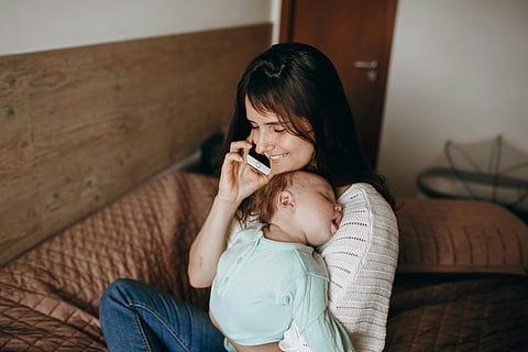 Smiling Mother Sitting with Sleeping Baby and Talking on Phone