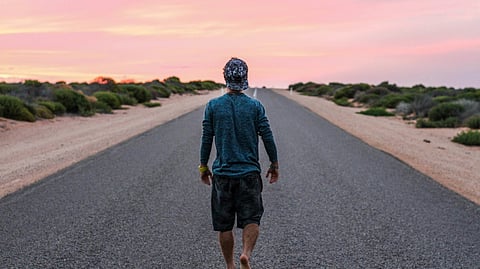 A man walking alone down the center of an empty road, surrounded by open space and clear skies.