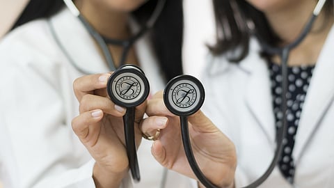 Two women in white coats holding stethoscopes, standing together in a clinical setting, smiling confidently.