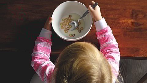 A young girl happily eats cereal from a bowl at a table, with a spoon in her hand and a smile on her face.