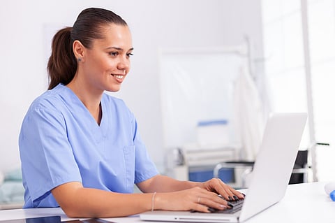 Healthcare worker in uniform using laptop sitting at desk in hospital office.