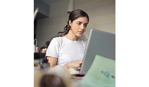 A woman seated at a desk, focused on her laptop, with a notepad and pen beside her.