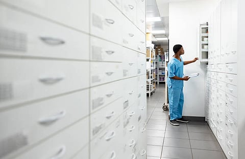 A man in scrubs stands in a pharmacy, surrounded by shelves filled with various medications and health products.