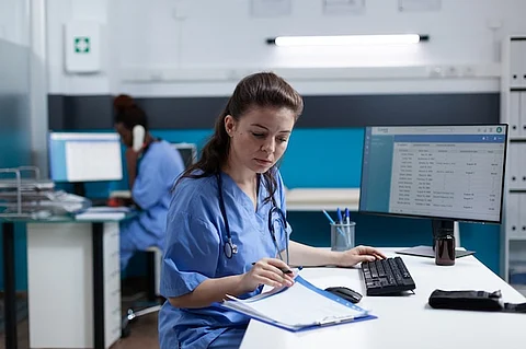 A female nurse in scrubs seated at a desk, focused on her work with medical tools and papers around her.