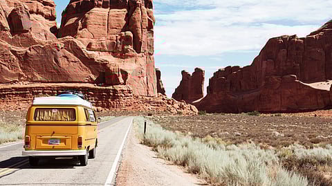 A yellow van is moving through a desert terrain with towering red rock formations on a two-lane road.