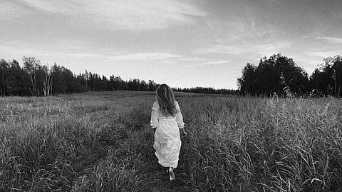 A Grayscale Photo of a Woman in White Dress Walking on Grass Field