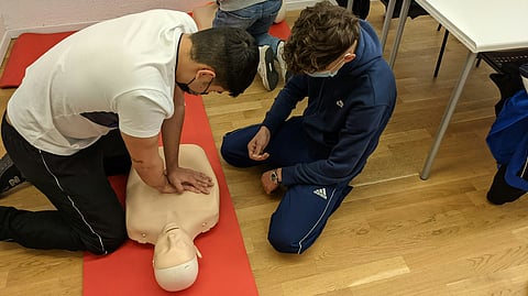 man in white t-shirt and blue pants sitting on floor and giving CPR to the dummy.