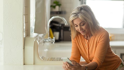 An older woman wearing an orange top standing in her kitchen browsing in her phone.