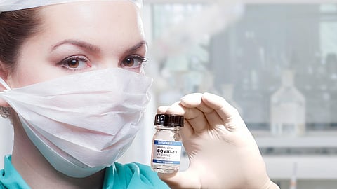 A healthcare worker, wearing protective gear including a face mask and gloves, holds a vial labeled "COVID-19 VACCINE".