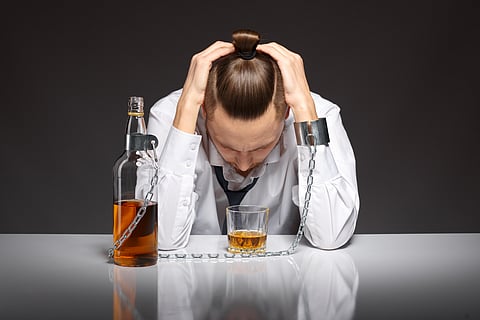 A man in a white shirt and black tie sits at a table with his head in his hands, handcuffed to an alcohol bottle.