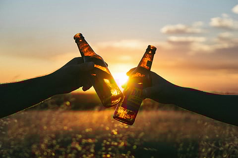 Two hands clinking beer bottle in an outdoor setup under sunset.