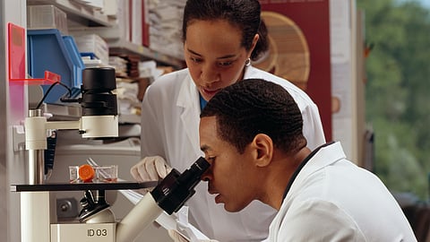 Two scientists wearing lab coat and gloves, one looking into the microscope while the other is guiding and assisting.