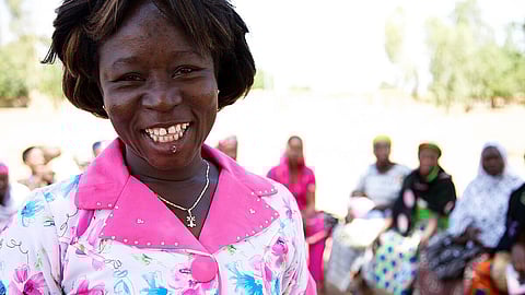 An African woman smiles in floral top with cross necklace, surrounded by others.