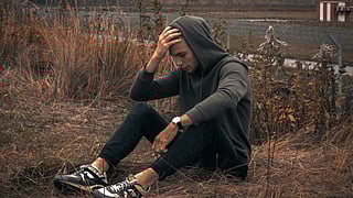 A man wearing a grey hoodie and black pants sitting outdoors on wilted grass by roadside holding his head.