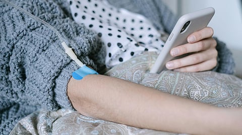 The female patient is scrolling through her phone while her right hand is resting because of IV drip.