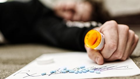 A blurred person lying with an outstretched hand holding an empty pill bottle with scattered pills nearby.