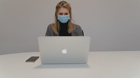 A woman wearing a mask is sitting in front of an open laptop with a mobile nearby.