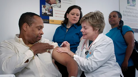 A female doctor administers an injection into the arm of a male patient, while two women are observing the process.