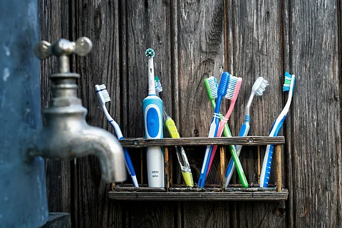 Toothbrushes, including one electric, stored in a wooden rack by a rustic wall with a metal water tap on the side.