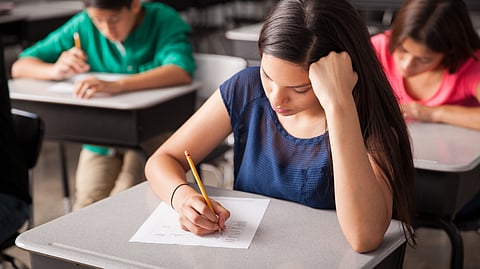 A young girl sitting with one hand supporting her head and another hand holding a pencil and filling OMR sheet.