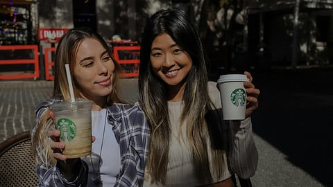Two girls holding Starbucks coffee in their hands and smiling at the camera.