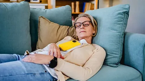 A woman resting with her eyes closed on couch, wearing casual attire, with a yellow phone amidst cozy cushions.