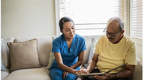 A nurse in blue scrubs helping a senior in a care facility to read some book.