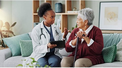 An elderly woman in a burgundy sweater and walking stick is sitting on a couch with a doctor in white lab coat.