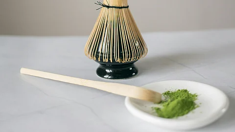 A green powder is kept on a white plate with a wooden spoon and a bamboo whisk is kept nearby.