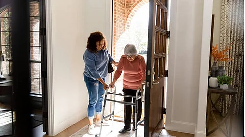 A young woman is helping an elder woman with a walking stick.