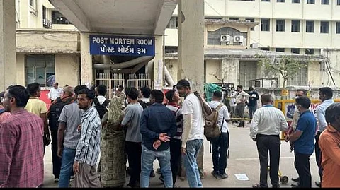 People gather in a large crowd outside the hospital's post-mortem wing, appearing to discuss or await information.