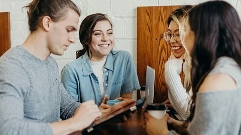 Four students sit at a wooden table in a cozy setting. They are engaged in conversation with laptops.