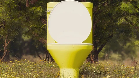 A bright yellow toilet stands in a field of flowers, surrounded by lush green trees.