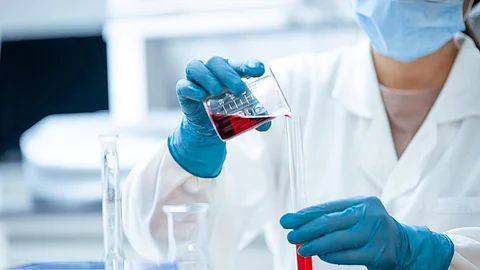 Scientist with gloves and mask pours red liquid from a beaker into a test tube in a lab.
